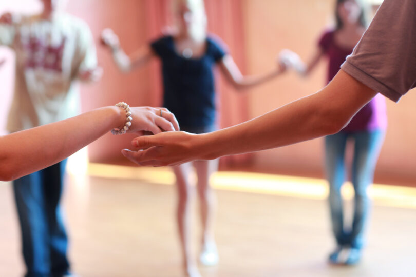 Nahaufnahme zweier junger Menschen, die sich bei einer Tanzübung an den Händen fassen, während weitere Personen unscharf im Hintergrund in einem hellen Raum ste