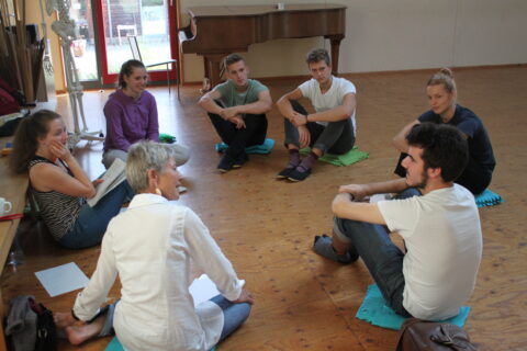 Eine Gruppe junger Erwachsener und eine ältere Frau sitzen barfuß im Kreis auf bunten Sitzmatten in einem hellen Raum mit Holzboden. Sie unterhalten sich entspa