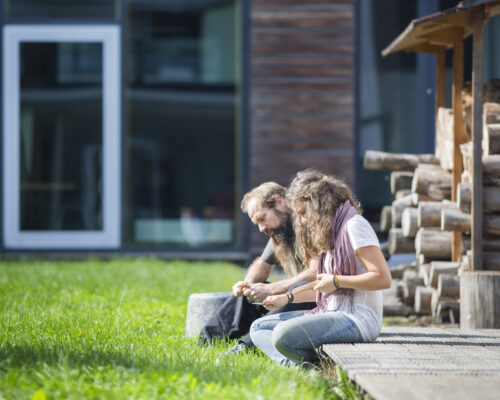 Zwei Personen sitzen draußen auf einer hölzernen Terrasse neben einem Holzstapel und schnitzen oder bearbeiten gemeinsam Holzstücke, im Hintergrund ein modernes