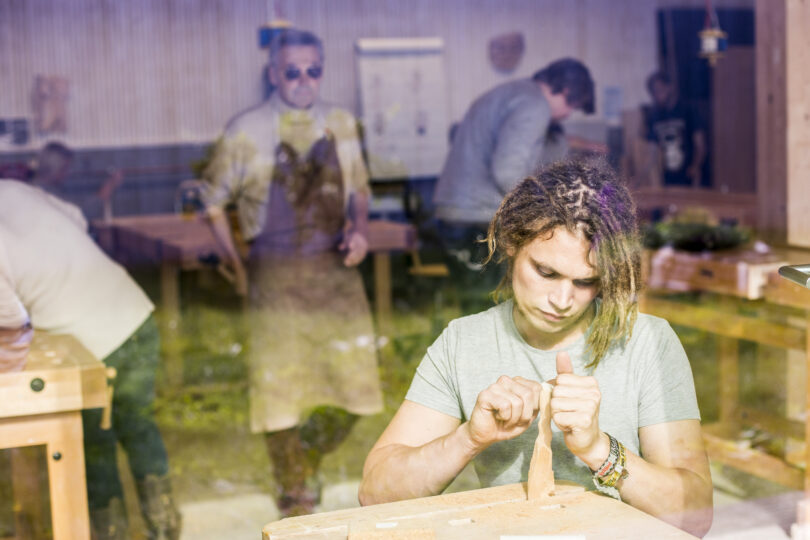Ein junger Mensch mit Dreadlocks sitzt konzentriert an einer Werkbank und schnitzt ein Holzstück, während im Hintergrund in einer hellen Holzwerkstatt mehrere a