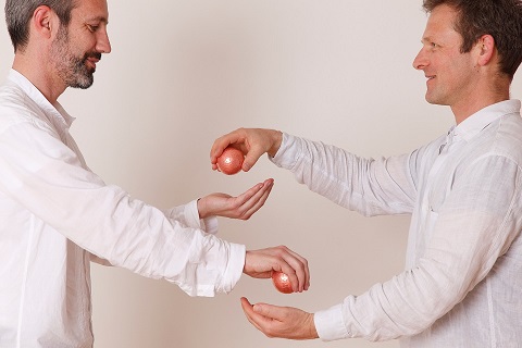 Zwei Männer in weißen Hemden stehen sich gegenüber und halten je eine kupferfarbene Kugel in den Händen, als würden sie diese achtsam übergeben oder eine Übung