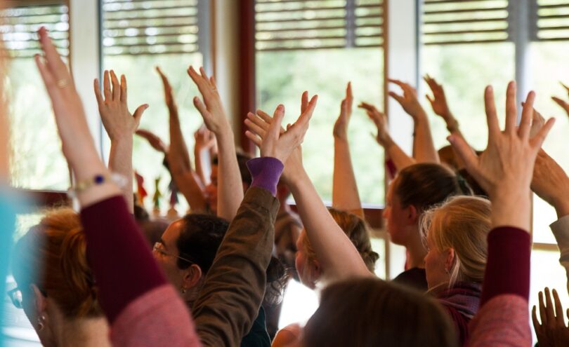 Eine Gruppe von Menschen sitzt in einem hellen Raum und streckt gleichzeitig beide Arme nach oben, als ob sie aktiv an einem Seminar oder Workshop teilnehmen.