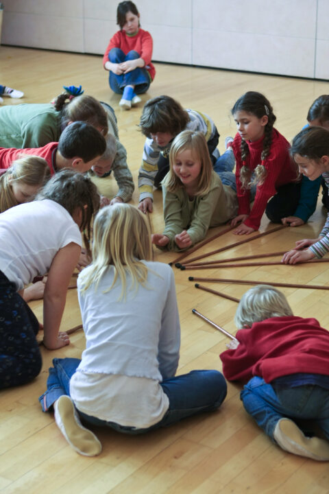 Eine Gruppe von Kindern sitzt und kniet im Kreis auf einem Holzfußboden und spielt gemeinsam mit mehreren langen Holzstäben, während ein weiteres Kind etwas abs