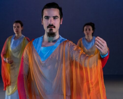 Zeitgenössische Tanzszene mit farbenfrohen, transparenten Kostümen Drei Tänzerinnen und Tänzer auf einer Bühne, die vordere Person mit Bart und Spitzbart trägt ein fließendes, orange-gelbes Übergewand über hellblauer Kleidung u