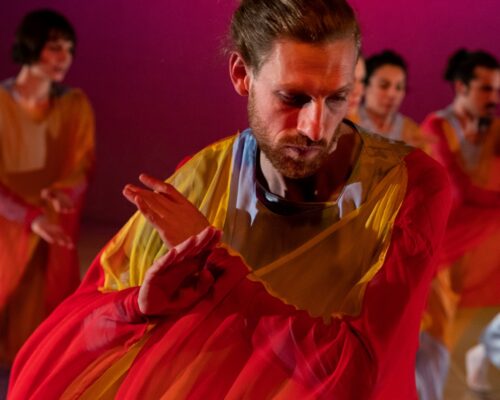 Zeitgenössische Tanzperformance in leuchtenden Farben Mehrere Personen tanzen auf einer Bühne vor einem pink beleuchteten Hintergrund. Im Vordergrund bewegt sich ein Mensch mit nach vorne geneigtem Kopf und gefalte