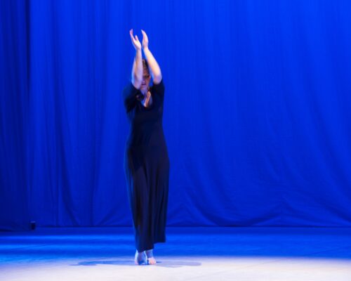 Solotanz auf blau beleuchteter Bühne Eine Person in einem langen, dunklen Kleid tanzt barfuß allein auf einer Bühne, die in intensivem Blaulicht beleuchtet ist. Die Arme sind über dem Kopf nach obe