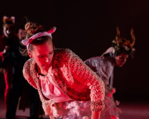 Theaterperformance mit fantasievollen Tierkostümen im roten Bühnenlicht Szene aus einer Theater- oder Tanzaufführung: Im Vordergrund beugt sich eine Darstellerin mit rosa Tierohren und flauschiger, rot angeleuchteter Jacke nach vorn