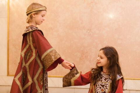 Zwei Kinder in prunkvollen mittelalterlichen Kostümen stehen sich gegenüber; das stehende Kind mit goldener Stirnbinde hält feierlich die Hand des sitzenden ode