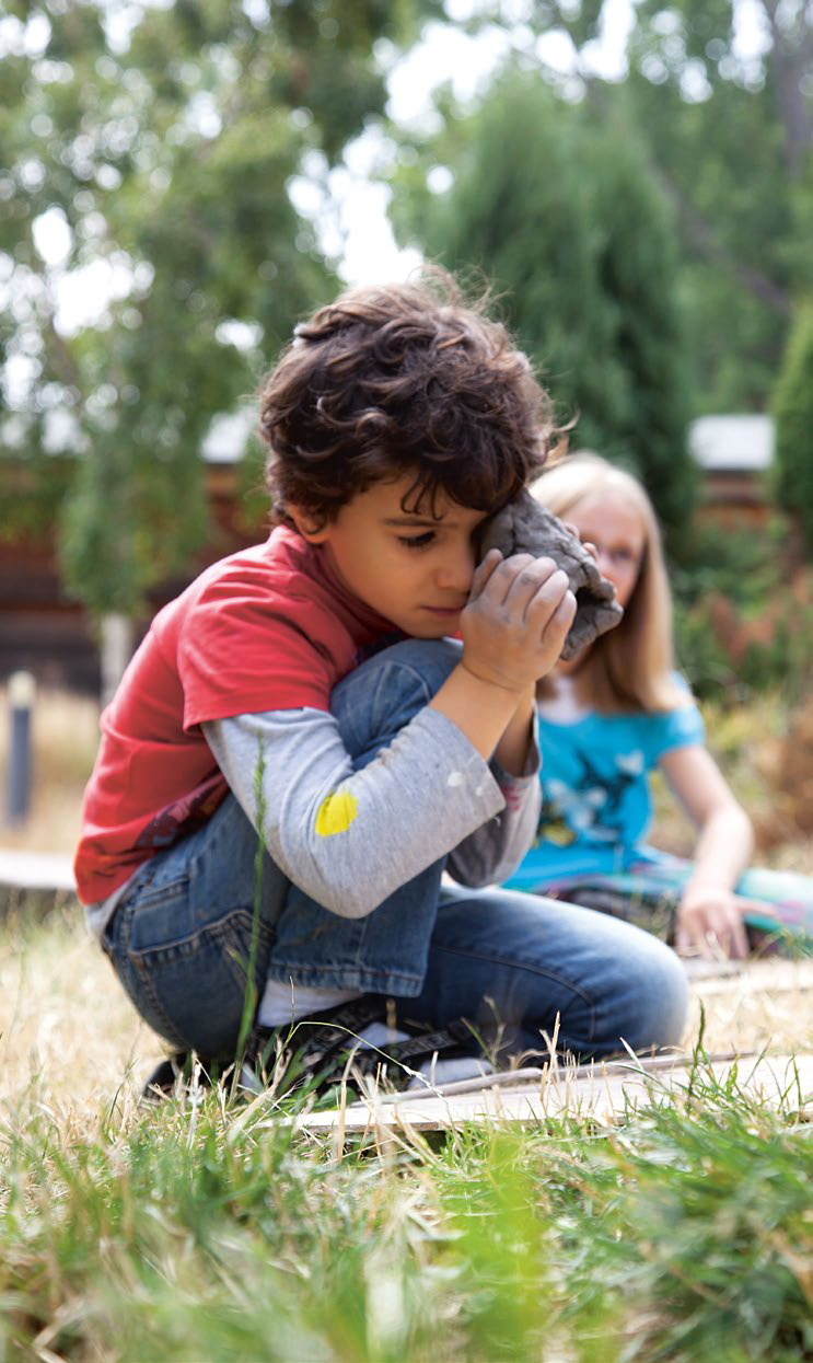 Kind mit lockigen Haaren hockt im Gras und hält ein Stück Ton an sein Ohr, während im Hintergrund ein weiteres Kind sitzt. Die Szene spielt im Freien zwischen B