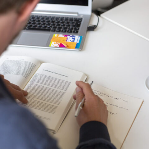 Eine Person sitzt an einem Schreibtisch, liest in einem aufgeschlagenen Buch und macht handschriftliche Notizen in ein Heft, im Hintergrund steht ein geöffneter