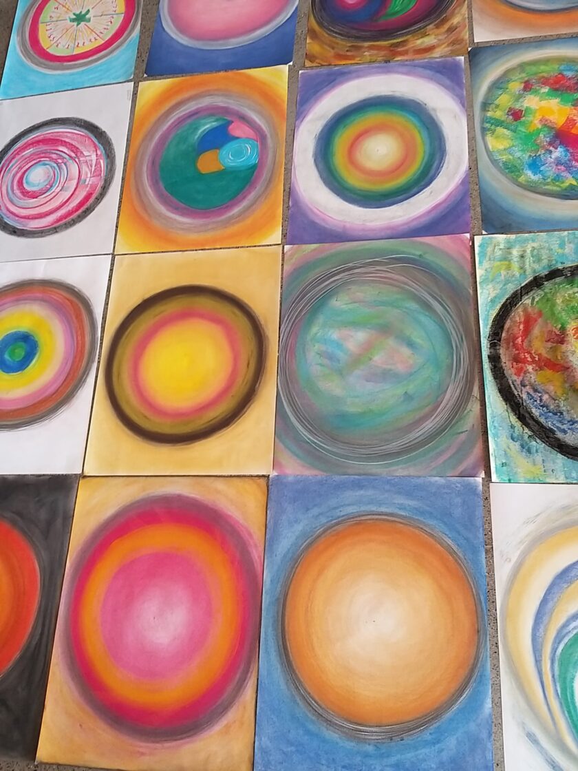 Mehrere rechteckige Papierblätter liegen auf dem Boden und zeigen bunte, kreisförmige Pastellzeichnungen. Jede Zeichnung besteht aus konzentrischen Ringen oder