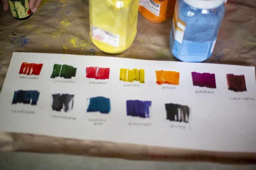 Nahaufnahme eines Blattes mit aufgemalten Farbfeldern in zwei Reihen, jeweils mit handschriftlich auf Deutsch beschrifteten Farbnamen wie „Persischrot“, „Lotusg