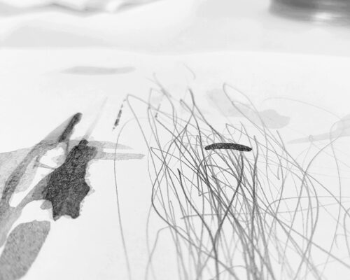 Abstrakte schwarz‑weiße Zeichnung auf Papier mit feinen, wirren Linien und einigen dunklen Tintenflecken, unscharfer Hintergrund mit Glas und Papieren auf einem