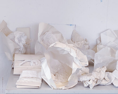 Ein grauer Tisch steht vor einer weißen Wand und ist mit zahlreichen zerknüllten und gefalteten weißen Papierbögen bedeckt, die wie eine abstrakte Papierlandsch