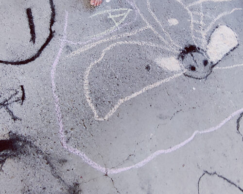 Kindliche Kreidezeichnung auf Asphalt mit einer großen, abstrakten Figur in Pastellfarben und einem Paar nackter Kinderfüße am oberen Bildrand.