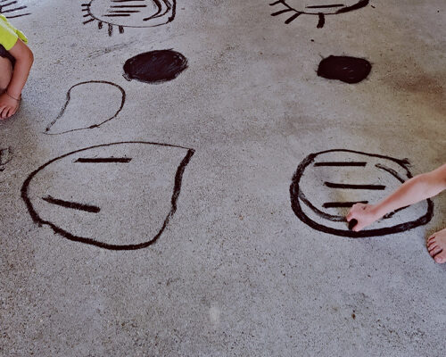 Zwei Kinder sitzen barfuß auf einem Betonboden und malen mit dunkler Farbe einfache Gesichter und Formen auf den Boden. Man sieht mehrere runde und ovale Zeichn