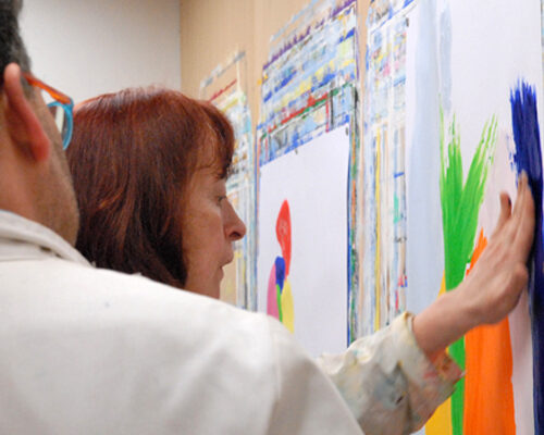 Zwei Personen in weißen Malkitteln stehen vor großen Papierbögen an der Wand; die Frau im Vordergrund verstreicht mit der Hand kräftige bunte Farben wie Blau, G