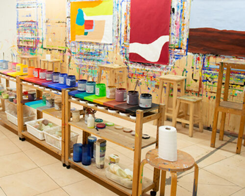 Helles Kunstatelier mit mehreren farbenfrohen abstrakten Gemälden an der mit Farbspritzern bedeckten Wand. Davor stehen zwei lange Holzwagen mit offenen Farbdos