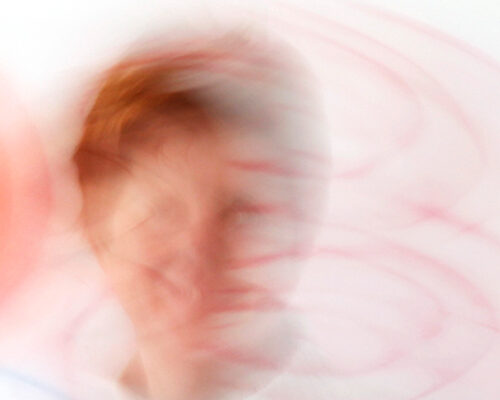 Stark verwischtes Porträt einer Person mit rötlichem Haar vor hellem Hintergrund, umgeben von wirbelnden rosa und roten Linien, die eine dynamische, abstrakte W