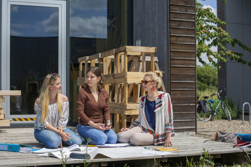 Drei junge Frauen sitzen draußen auf einer Holzterrasse vor einem modernen Gebäude und arbeiten gemeinsam an großen Papierbögen und Unterlagen, die auf dem Bode