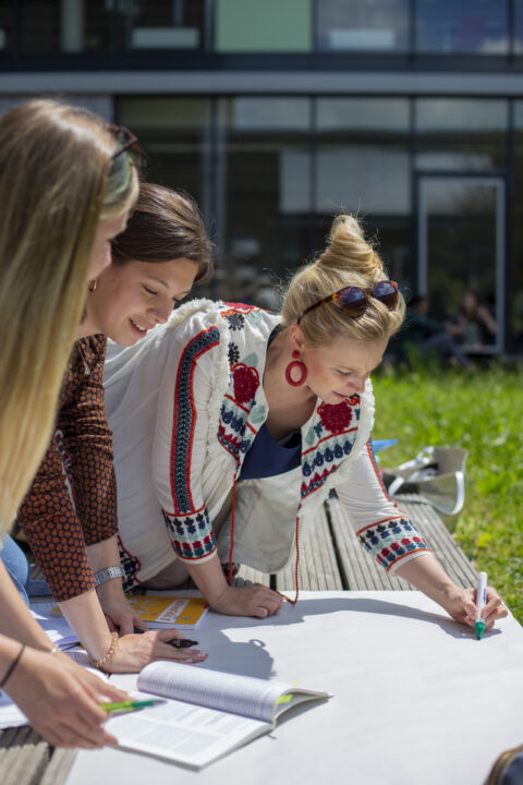 Drei junge Frauen arbeiten gemeinsam im Freien an einem großen weißen Plakat, beugen sich darüber und schreiben Notizen, während Bücher und Unterlagen vor ihnen