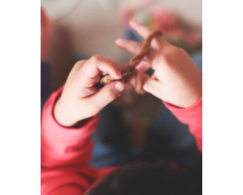 Nahaufnahme von Kinderhänden in einem pinken Langarmshirt, die einen braunen Faden oder eine Schnur zwischen den Fingern halten und damit spielen.