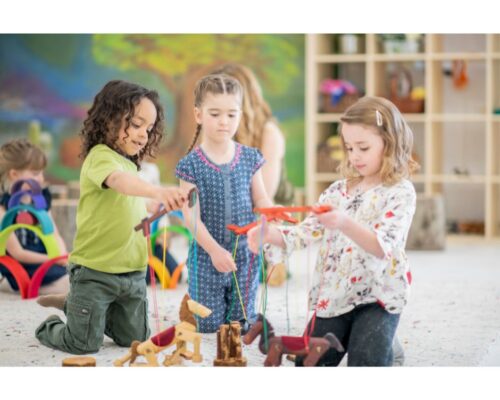 Drei Kinder spielen gemeinsam auf dem Boden einer Kita mit hölzernen Pferde-Marionetten, während im Hintergrund weitere Kinder mit bunten Holzspielzeugen beschä