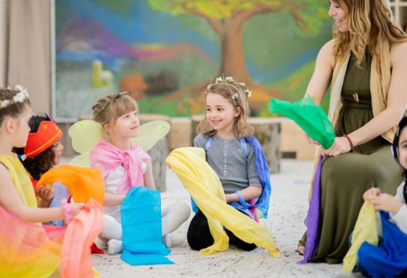 Gruppe von Kindern sitzt im Kreis auf dem Boden in einem bunt dekorierten Raum und spielt mit farbigen Tüchern, während eine Erzieherin mit ihnen lacht und mitm