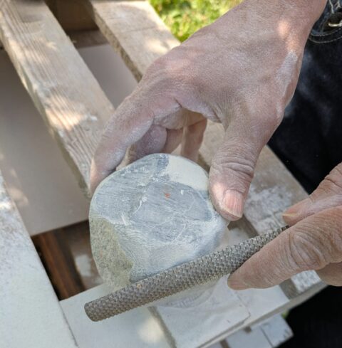 Nahaufnahme von staubigen Händen, die mit einer rauen Feile einen hellen Stein bearbeiten, Holzoberfläche im Hintergrund.