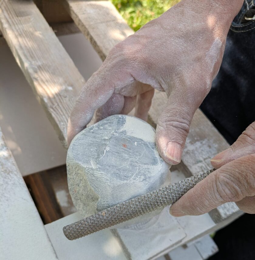 Nahaufnahme von staubigen Händen, die mit einer rauen Feile einen hellen Stein bearbeiten, Holzoberfläche im Hintergrund.