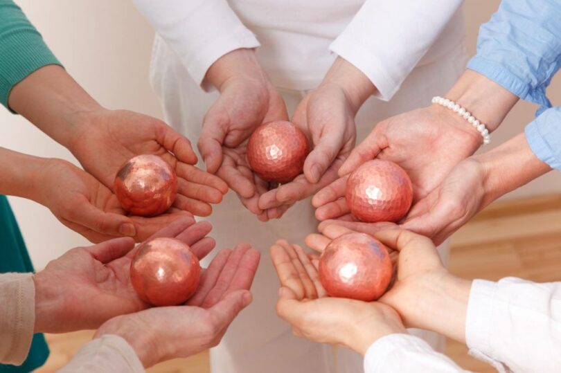 Mehrere Personen stehen im Kreis und halten in ihren geöffneten Händen glänzende, gehämmerte Kupferkugeln, die sie einander zuwenden.