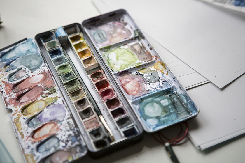 Offenes Aquarellkasten-Set mit vielen bereits benutzten, bunt verlaufenen Farbnäpfen und zwei Pinseln in der Mitte, daneben liegen weiße Zeichenpapiere auf eine