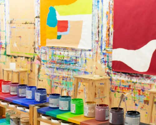 Atelier mit vielen offenen Farbdosen in Regenbogenreihenfolge auf einem Holztisch; im Hintergrund hängen großformatige abstrakte Bilder auf farbbesprenkelten Wä