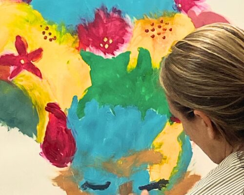 Person mit nach hinten gebundenen Haaren steht vor einer Leinwand und malt mit den Fingern an einem bunten, abstrakten Bild mit kräftigen Farben wie Blau, Grün,