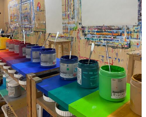 Reihe offener Farbdosen in Regenbogenfarben auf einem Arbeitstisch in einem Atelier, jede Dose mit einem Metallspachtel, dahinter leere Blätter und eine bunt be