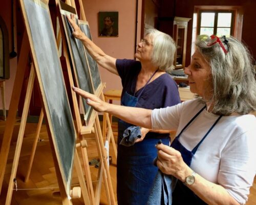 Zwei Frauen beim Malen im Atelier Zwei ältere Frauen stehen nebeneinander in einem Atelier und arbeiten konzentriert an Bildern auf Staffeleien. Beide tragen dunkle Schürzen und halten Tücher in