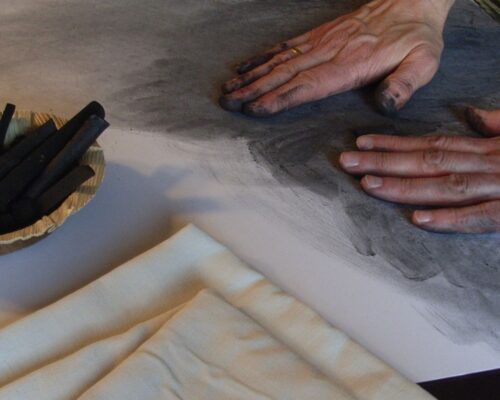 Mit Kohle und Händen zeichnen Zwei Hände verwischen schwarzen Zeichenkohlestaub auf einem Blatt Papier, daneben liegen ein Stapel heller Tücher und eine Schale mit Kohlestiften.