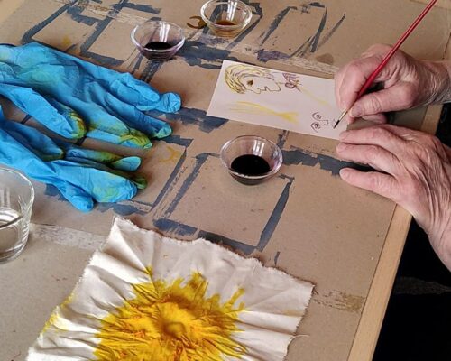 Eine Person malt mit einem feinen Pinsel auf einem Blatt Papier ein Porträt, daneben liegen blaue Einweghandschuhe, kleine Schälchen mit flüssiger Farbe, ein Gl