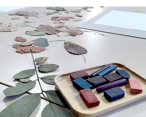 Auf einem hellen Tisch liegen zahlreiche getrocknete Blätter in verschiedenen Formen und Herbstfarben ausgebreitet. Im Vordergrund steht ein rechteckiges Holzta