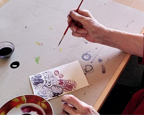 Eine ältere Person sitzt an einem Tisch und malt mit einem feinen Pinsel ein abstraktes Motiv in Lila- und Rottönen auf eine kleine Karte. Auf dem Basteltisch l