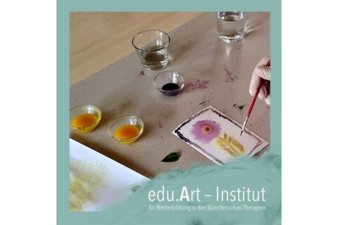 Eine Hand mit Pinsel malt auf einer kleinen Karte eine lila Blume und gelbe Blätter. Daneben stehen mehrere Glasschälchen mit farbiger Flüssigkeit und zwei Wass