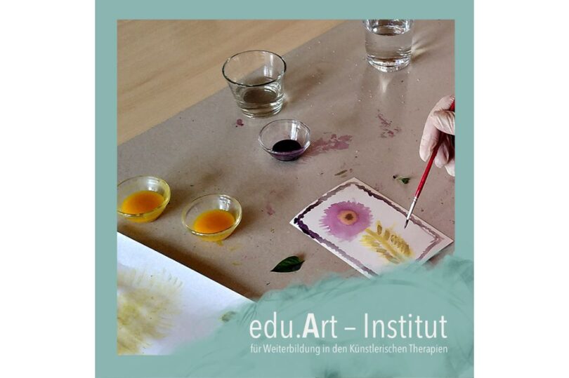 Eine Hand mit Pinsel malt auf einer kleinen Karte eine lila Blume und gelbe Blätter. Daneben stehen mehrere Glasschälchen mit farbiger Flüssigkeit und zwei Wass