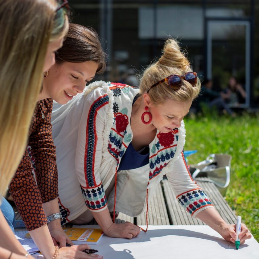 Drei junge Frauen arbeiten gemeinsam im Freien an einem großen weißen Plakat; eine Frau beugt sich vor und schreibt mit einem grünen Marker darauf, die anderen