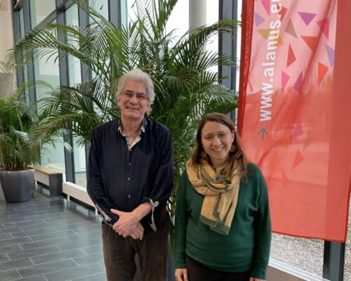 Zwei Personen stehen lächelnd in einem modernen Foyer mit großen Fenstern und vielen grünen Palmen. Links steht ein älterer Mann mit grauen Haaren und dunklem H