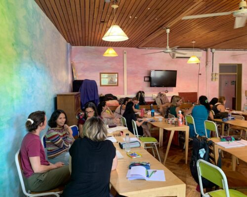 Mehrere Gruppen von Erwachsenen sitzen in einem farbenfroh gestrichenen Seminarraum an Holztischen und diskutieren miteinander. Auf den Tischen liegen Notizbüch