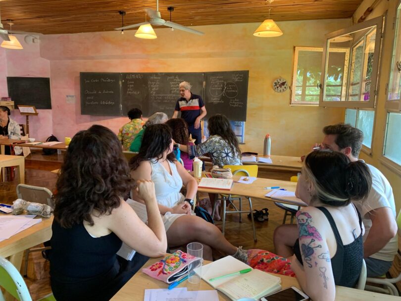 Gruppe von Erwachsenen sitzt in einem hellen Klassenraum an Holztischen, hört einem Lehrenden vorne an der Tafel zu und macht sich Notizen. Auf den großen Kreid