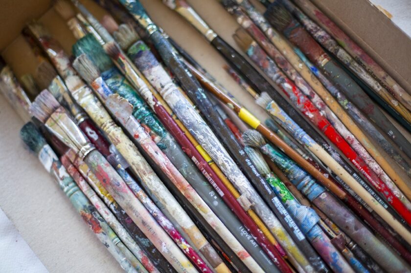 Nahaufnahme einer Schachtel voller unterschiedlich großer, stark benutzter Malpinsel, deren Stiele und Borsten mit bunten Farbresten bedeckt sind.