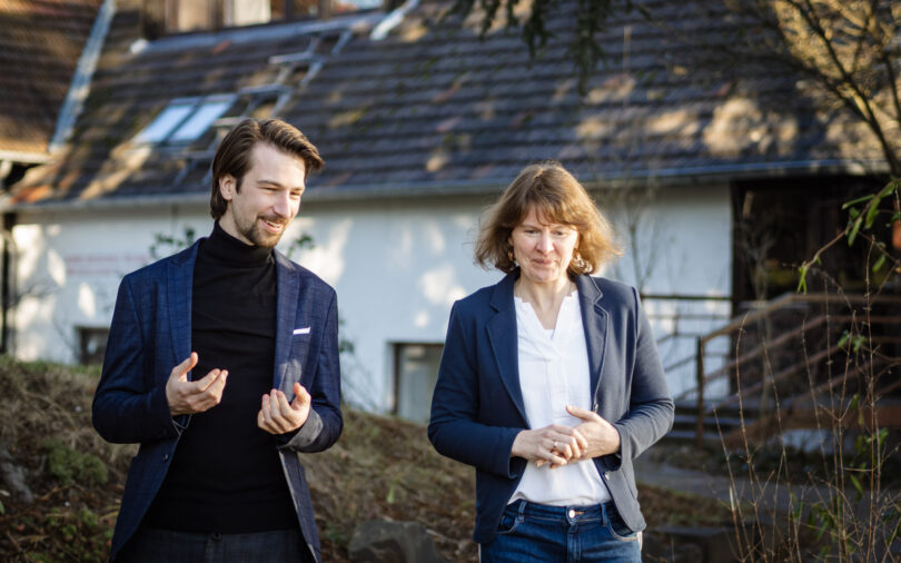 Zwei Menschen im Business-Look gehen nebeneinander im Freien und unterhalten sich, im Hintergrund ist ein Haus mit Schrägdach zu sehen.