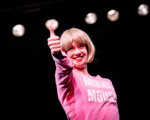 Lächelnde Person mit blondem Bob-Haarschnitt und pinkem Pullover mit der Aufschrift „Minnie Mouse“ zeigt mit nach vorn gestrecktem Arm den Daumen nach oben, vor