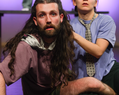 Zwei Personen sitzen auf einer Theaterbühne und blicken intensiv nach vorne. Die vordere Person mit langem, lockigem Haar und Bart stützt sich mit einer Hand au